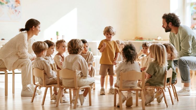 Kind steht mit erhobener Hand in der Mitte eines Stuhlkreises, während zwei Fachkräfte und die anderen Kinder es ansehen
